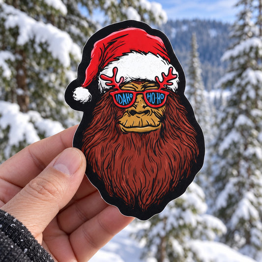 Idaho-Ho-Ho Bigfoot Santa sticker held outdoors in a snowy Idaho forest